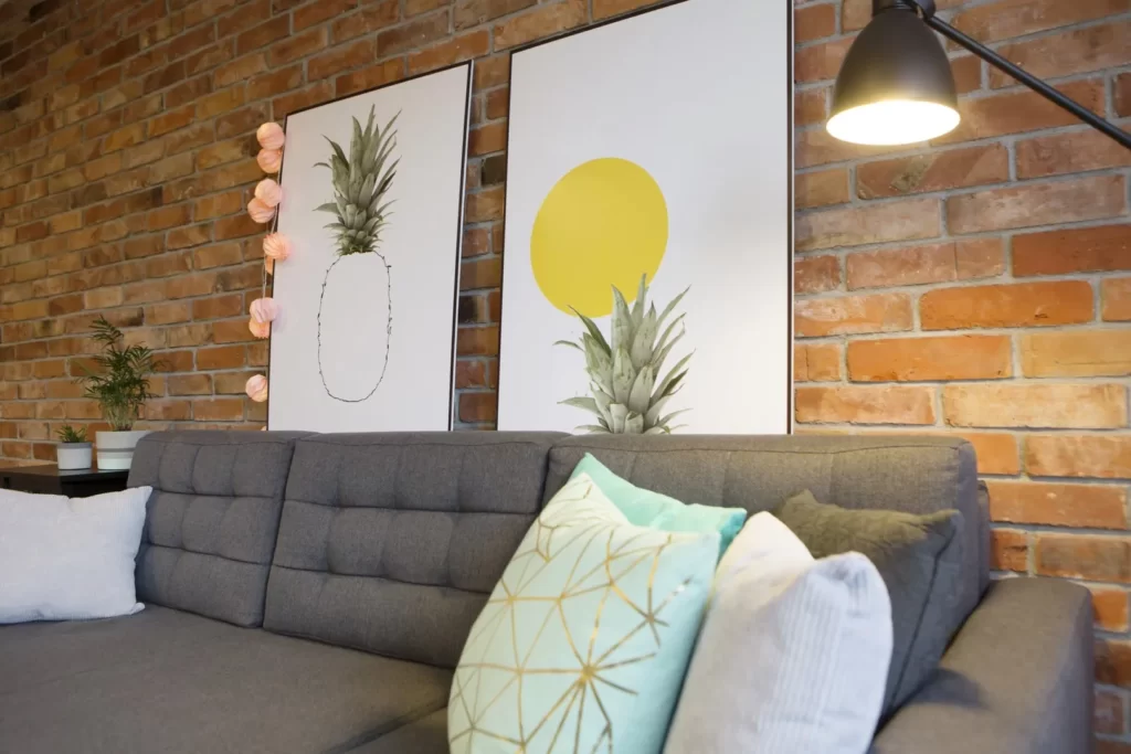 Nowoczesny kącik w salonie z szarą sofą, dekoracyjnymi poduszkami i dziełami sztuki na tle ceglanej ściany.