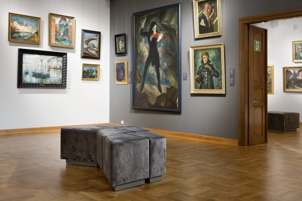 Wnętrze galerii sztuki z obrazami na ścianach i ławką w środku.