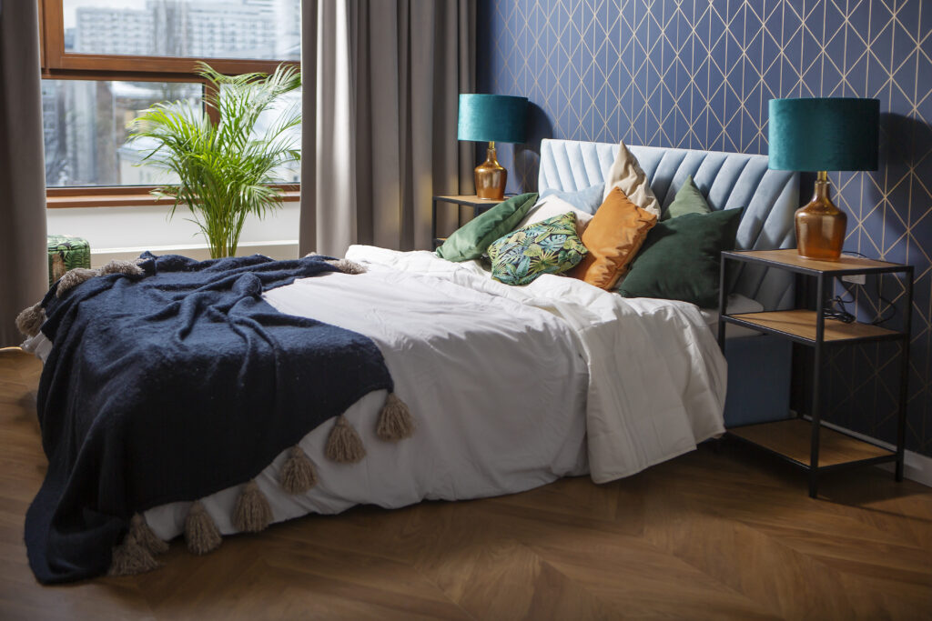 Elegancka sypialnia z niebieskim wezgłowiem łóżka, wzorzystą tapetą i kolorowymi poduszkami.