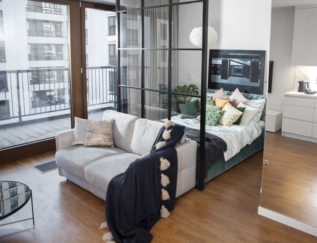 Nowoczesny salon z kanapą , szklanym stolikiem kawowym i widokiem na sypialnię przez szklaną ściankę działową.