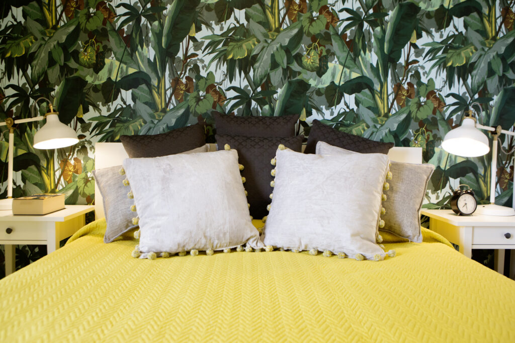 Przytulna sypialnia, lóżko z żółtą narzutą, tapeta z motywem roślinnym i białymi stolikami nocnymi.