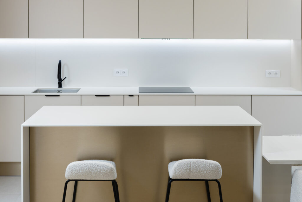 Nowoczesna, minimalistyczna kuchnia z eleganckimi, białymi szafkami i dwoma miękkimi krzesłami przy wyspie.